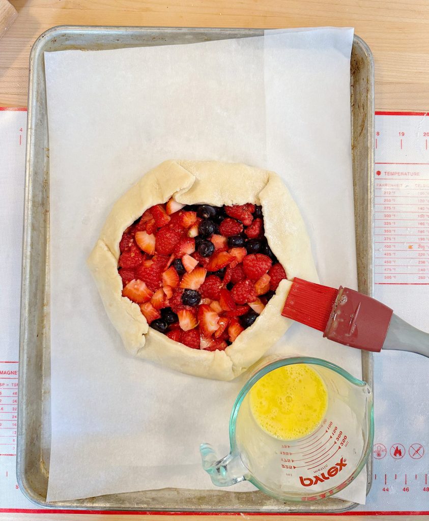 Unbaked berry galette on a baking sheet with parchment paper, beside a glass measuring cup with an egg wash and a pastry brush.