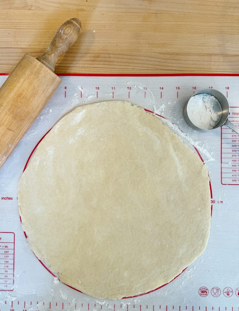 Rolled out pie dough on a measuring mat with a rolling pin and dough scraper nearby, preparing for baking.
