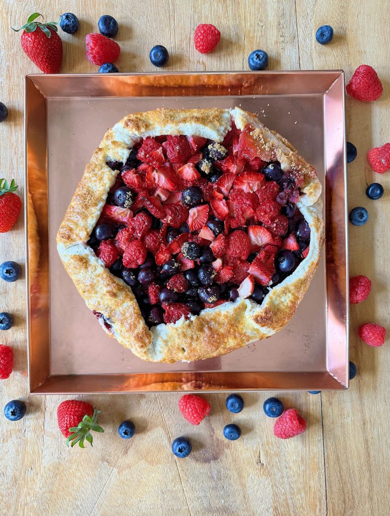 Rustic berry galette with strawberries, raspberries, and blueberries on a copper tray, surrounded by scattered berries on a wooden surface.