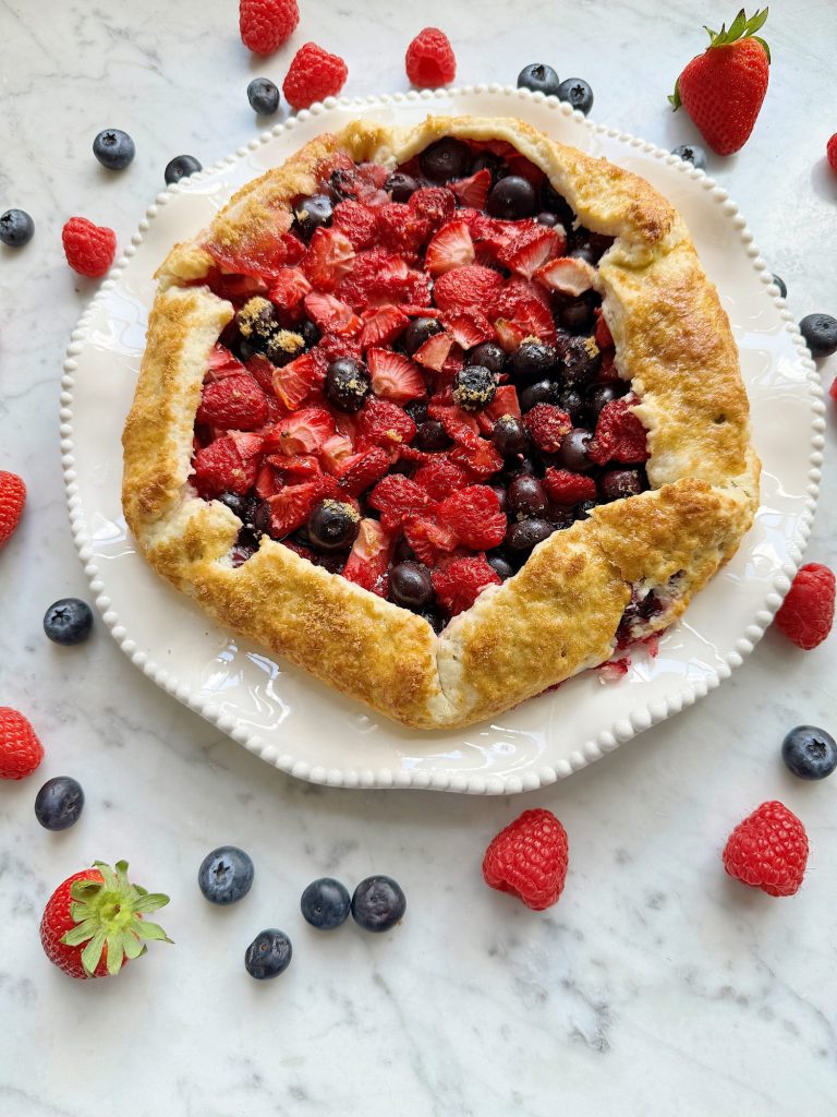 A freshly baked mixed berry galette with strawberries and blueberries on a decorative plate, surrounded by scattered berries on a marble surface.