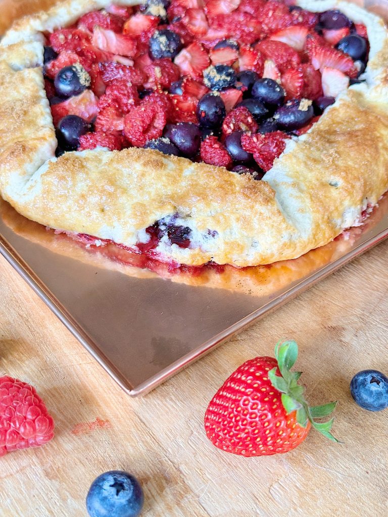 Mixed berry galette with a golden crust on a wooden board, garnished with fresh strawberries, raspberries, and blueberries.