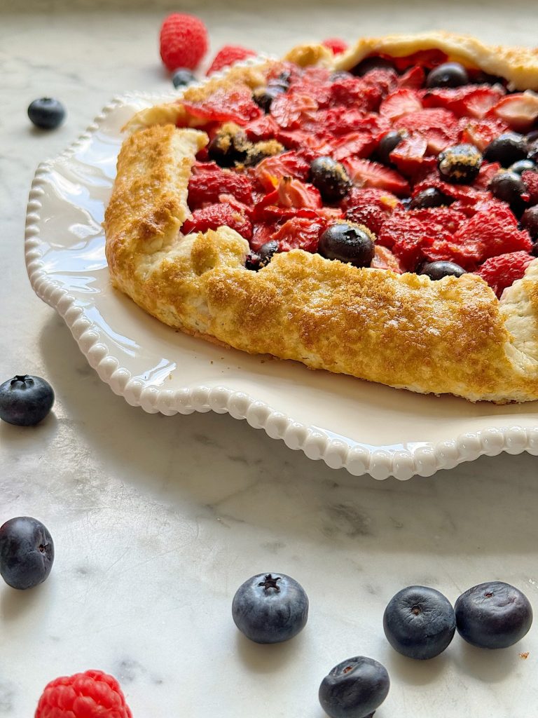 A freshly baked strawberry and blueberry galette on a white plate, surrounded by scattered blueberries and raspberries on a marble surface.