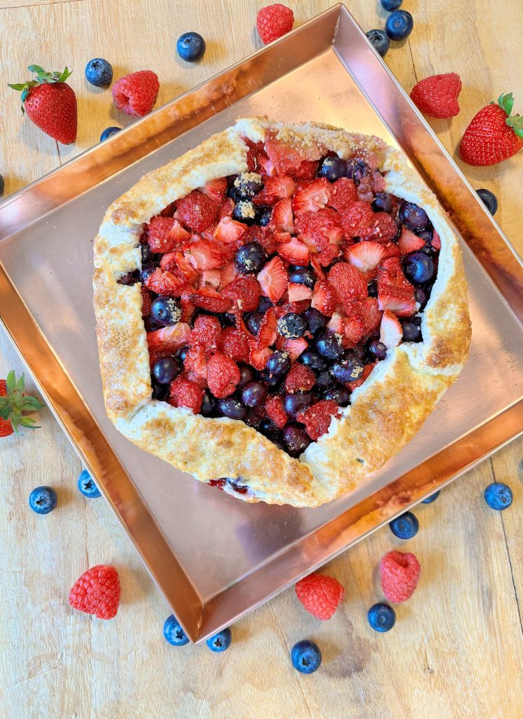 Freshly baked berry galette with strawberries, blueberries, and raspberries on a copper tray surrounded by scattered berries.