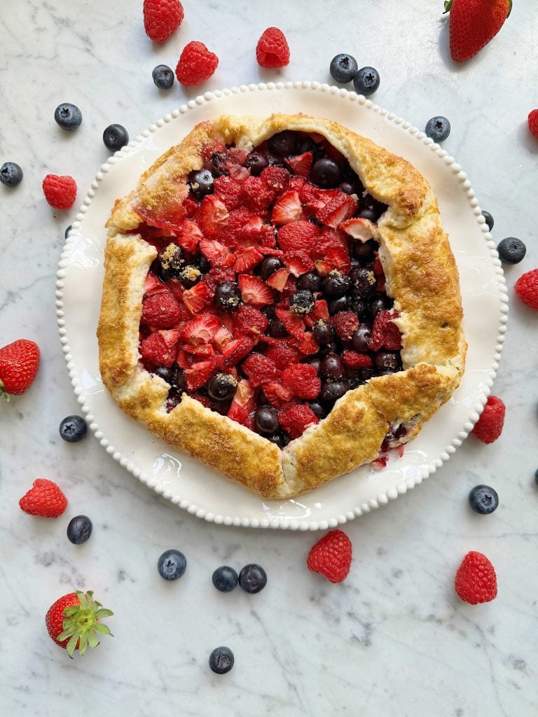 A fresh fruit galette with strawberries, raspberries, and blueberries on a white plate, with scattered berries in the foreground on a marble surface.
