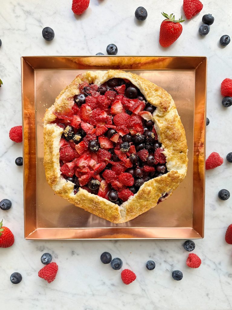 Freshly baked berry galette with strawberries, blueberries, and raspberries on a copper tray surrounded by scattered berries.