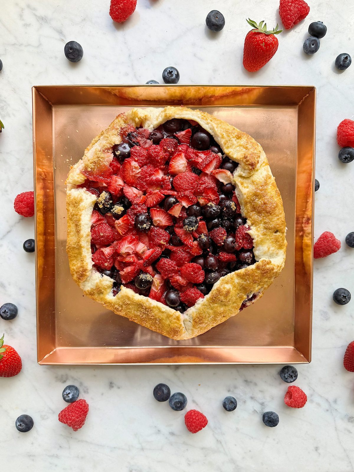 Freshly baked berry galette with strawberries, blueberries, and raspberries on a copper tray surrounded by scattered berries.