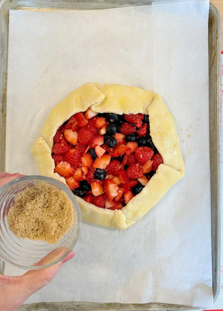 Unbaked mixed berry galette on a baking sheet, with a hand sprinkling brown sugar over the fruit filling.