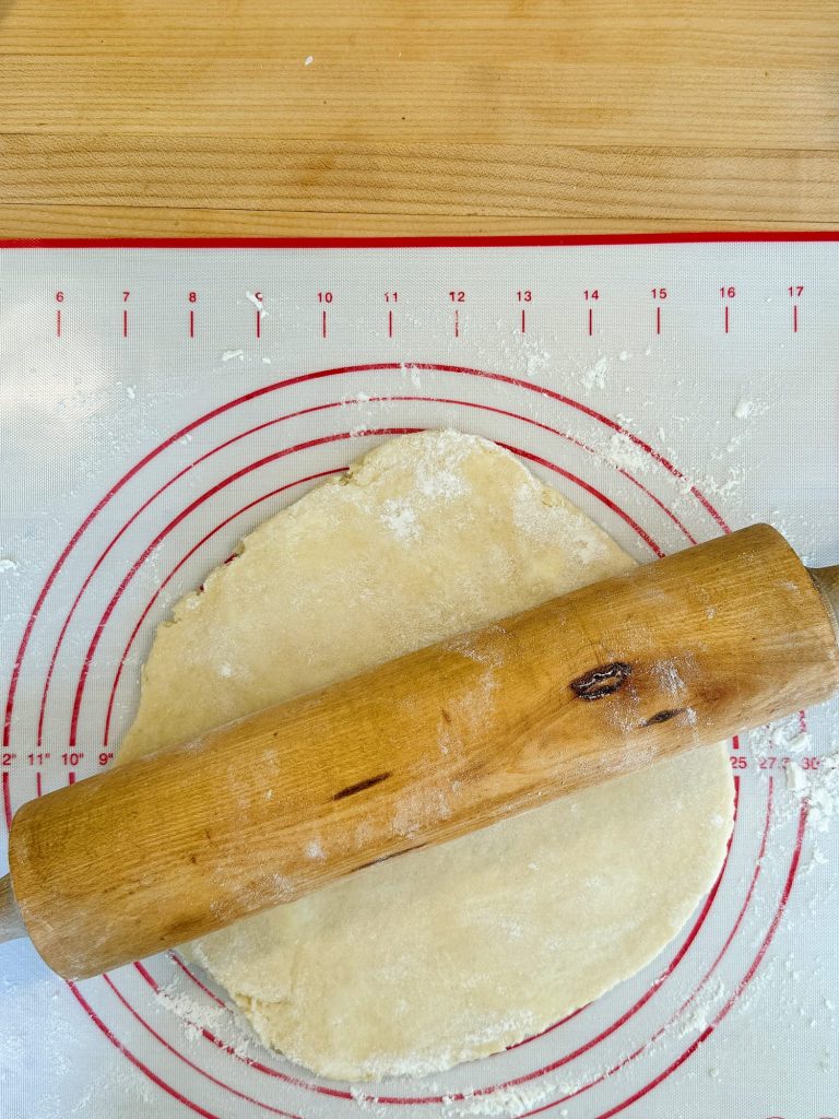 Rolled-out dough on a flour-dusted silicone mat with printed measurements, accompanied by a wooden rolling pin.