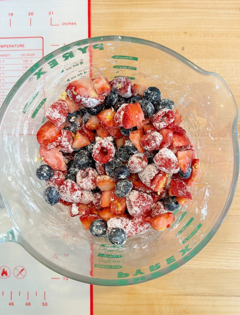 Glass bowl containing strawberries, raspberries, and blueberries sprinkled with sugar, resting on a measuring mat with temperature and size guides.