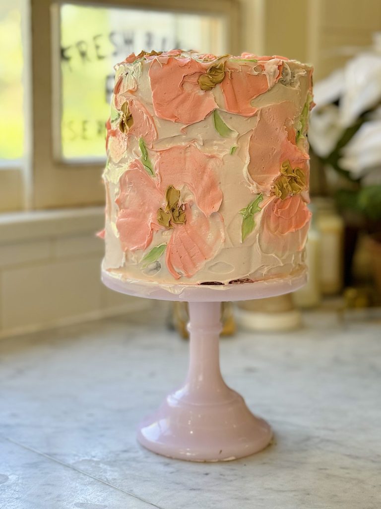 A tall, multi-layer cake adorned with pink buttercream and peach-colored floral decorations, displayed on a pink cake stand in a sunny kitchen.