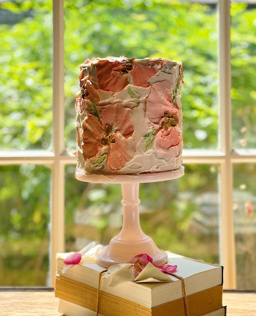 A tall, multilayered cake with pink and brown frosting, decorated with flowers, displayed on a pink cake stand on top of stacked books, in front of a window overlooking a garden.