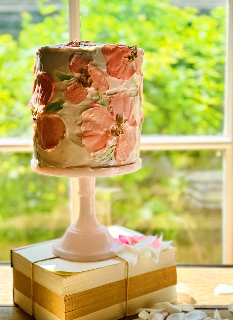 A tall, frosted cake with pink floral decorations, displayed on a cake stand beside a window overlooking greenery.