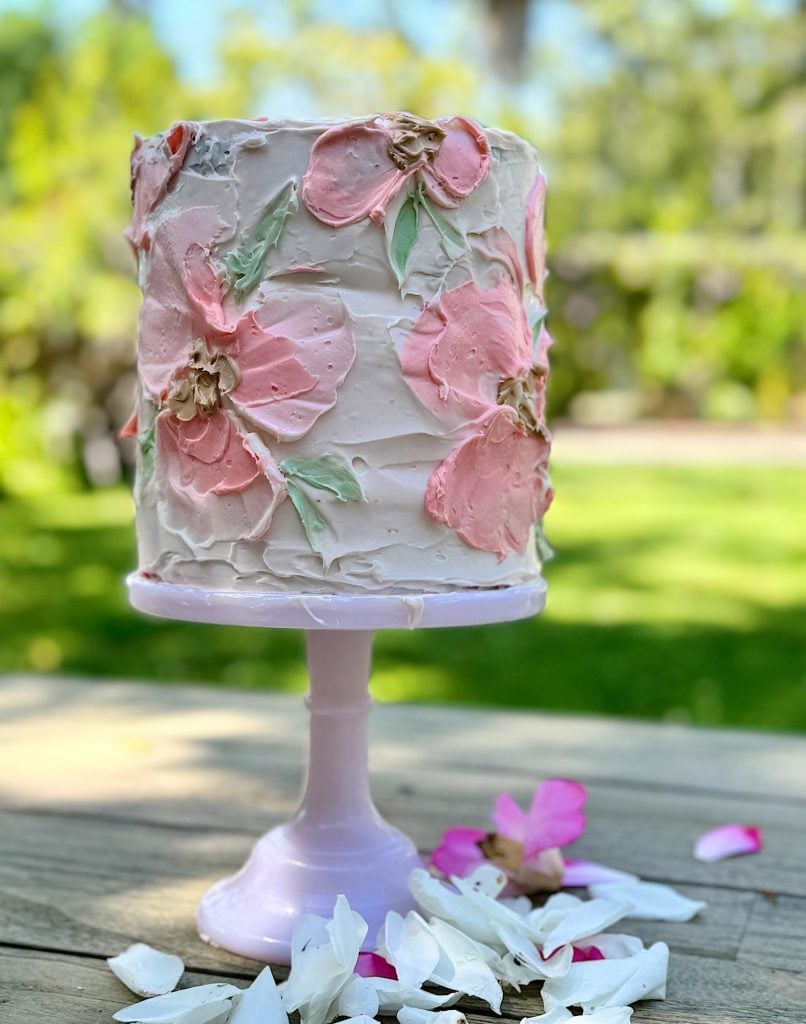 Tall, decorated cake with pink floral frosting patterns on a purple stand, surrounded by white petals, outdoors.