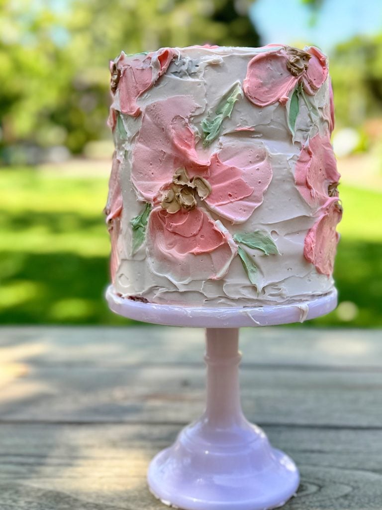 A tall, layered cake with pink and white floral frosting designs, displayed on a pink cake stand outdoors.