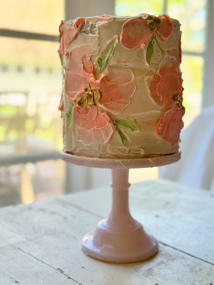 A tall, multi-layered cake with blush and orange floral frosting designs on a pink cake stand, displayed near a window with natural light.