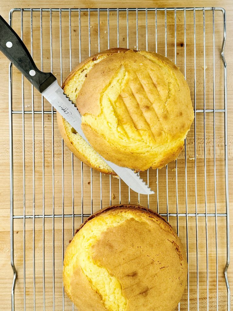 Two freshly baked round cakes on a cooling rack, one being sliced by a knife.