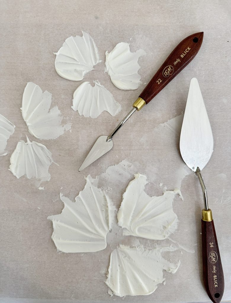 Leaves-shaped white frosting and two cake decorating tools, a palette knife and a sculpting tool, on a marble surface.