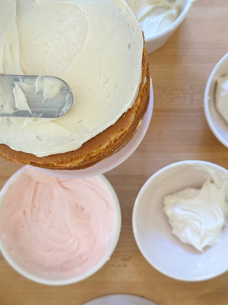 Top view of a person frosting a cake with creamy white icing, with bowls of pink and white frosting on a wooden table.