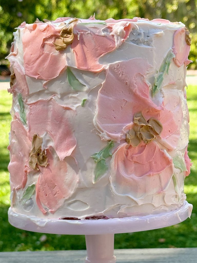 A tall cake with textured white and pink frosting and painted flowers, displayed outdoors.