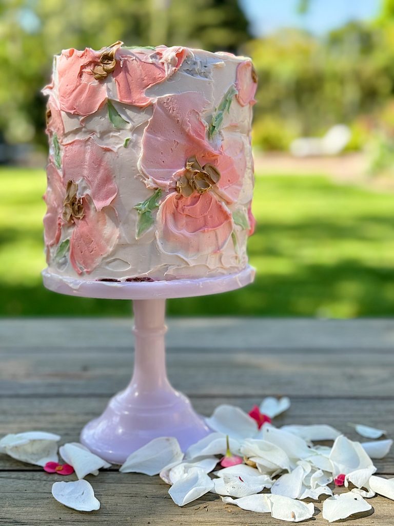 A tall, multi-layered cake with pink and white frosting flowers on a pink cake stand, surrounded by scattered petals on a wooden table outdoors.