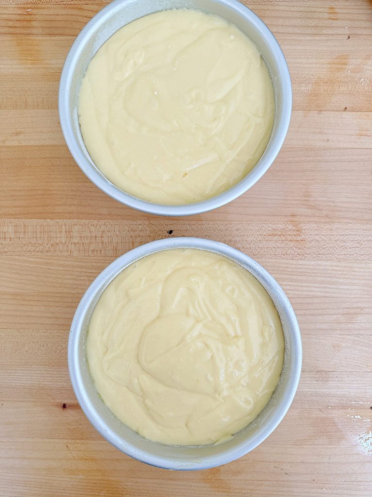 Two round cake pans filled with uncooked cake batter on a wooden table.
