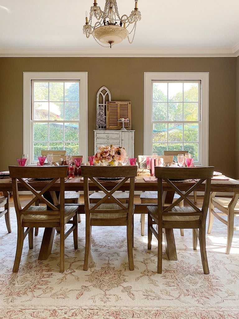 Elegantly set dining table in a room with neutral walls and large windows, featuring wooden chairs and a decorative centerpiece.