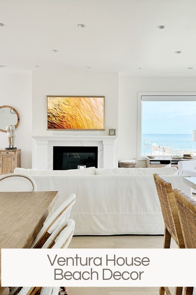 Modern beach house living room with white walls, a mounted tv above a fireplace, wooden dining table, and a view of the ocean through large windows.