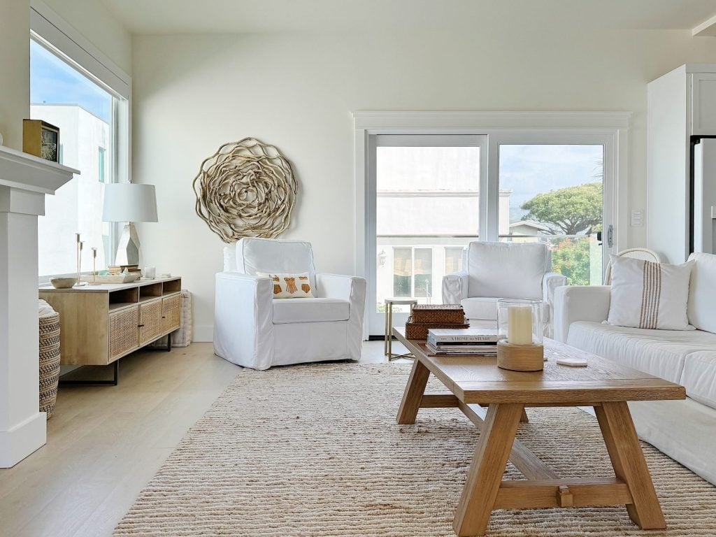 Bright, modern living room with white sofas, wooden coffee table, large textured wall art, and windows showing exterior view.
