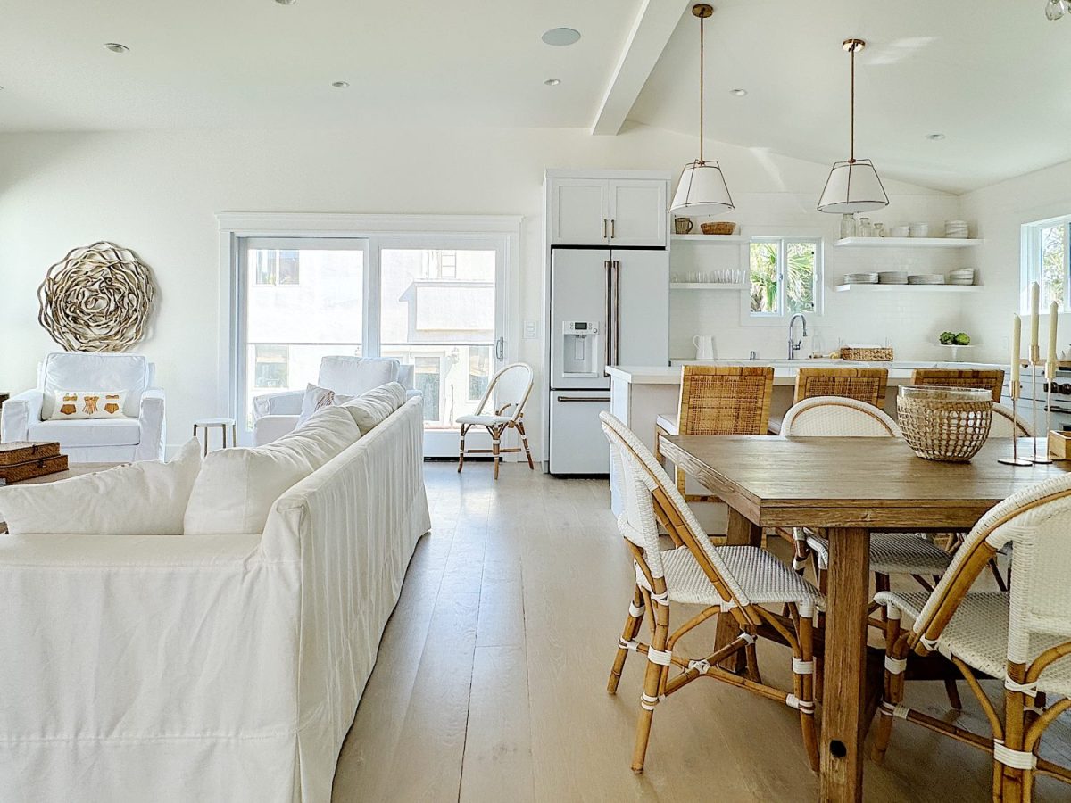 Bright, open-plan kitchen and living room with white decor, wooden dining table, rattan chairs, and modern appliances.