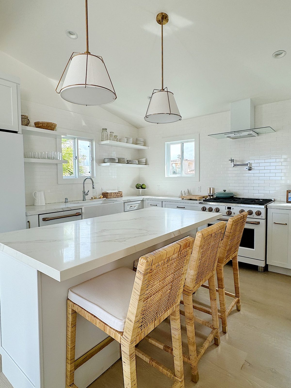 Bright modern kitchen with white cabinetry, floating shelves, a central island with a marble countertop, and rattan bar stools under pendant lights.