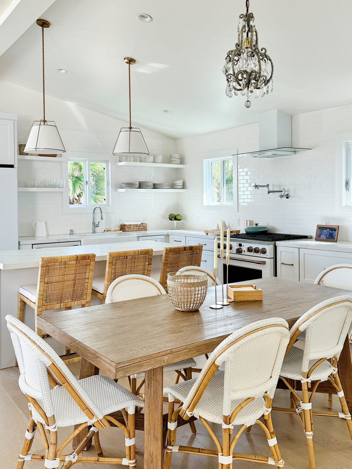 Bright, modern kitchen with white cabinetry, a wooden dining table, rattan chairs, hanging pendant lights, and chandelier.