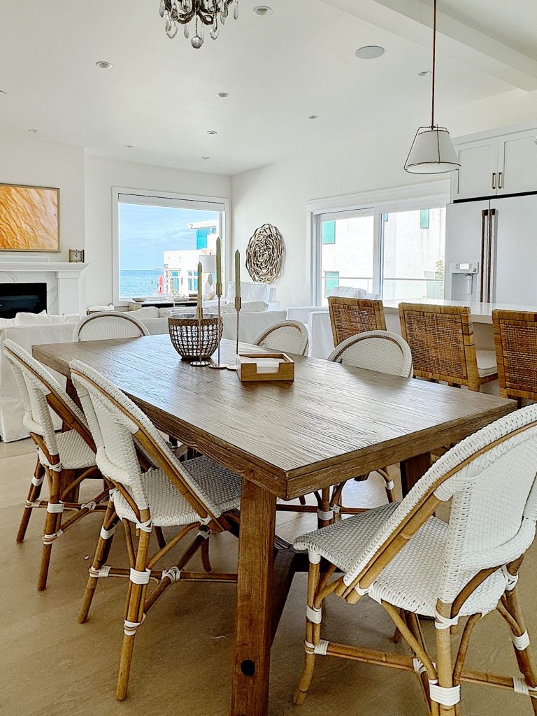 A bright dining room with a rustic wooden table, white wicker chairs, and a view of the sea through windows. modern kitchen visible in the background.