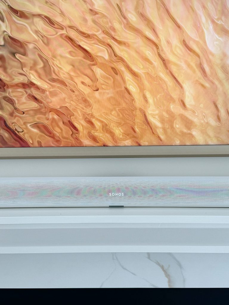 A wall-mounted sonos speaker on a white ledge against a textured orange and creamy abstract painting.