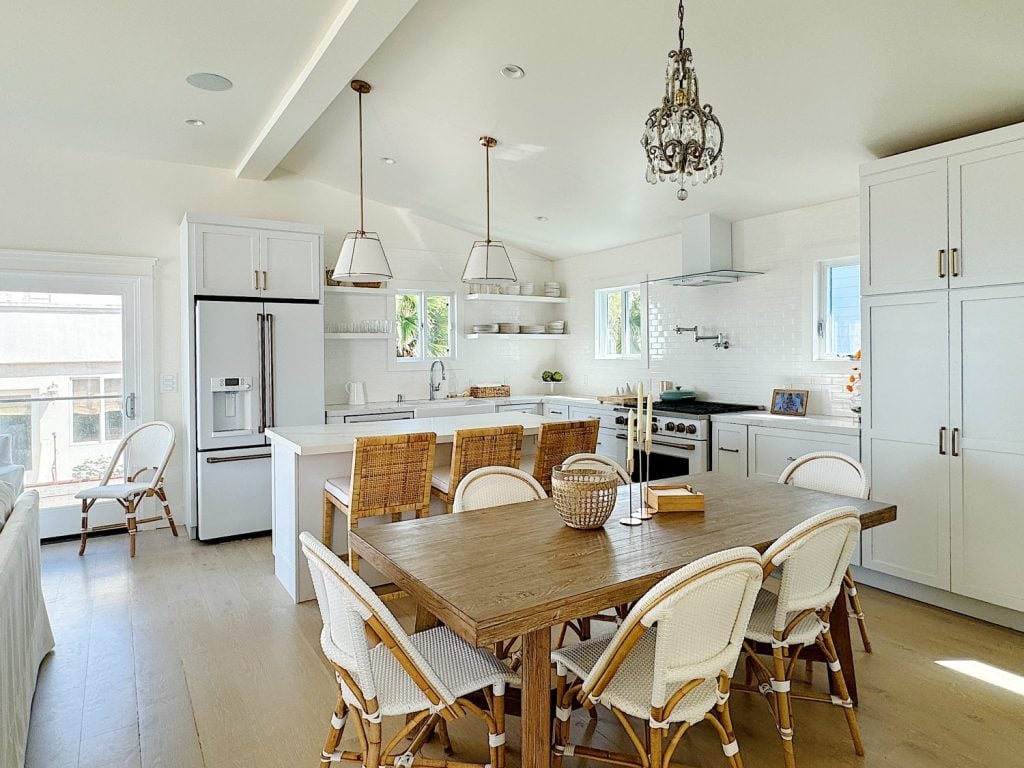 Bright, modern kitchen with white cabinets, stainless steel appliances, a wooden dining table with wicker chairs, and pendant lighting.