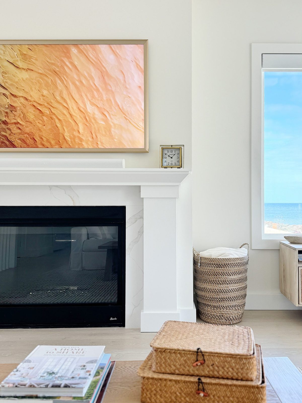 Modern living room with a fireplace, above which hangs a large abstract painting. a window to the right offers a view of the ocean. wicker baskets and magazines are in the foreground.