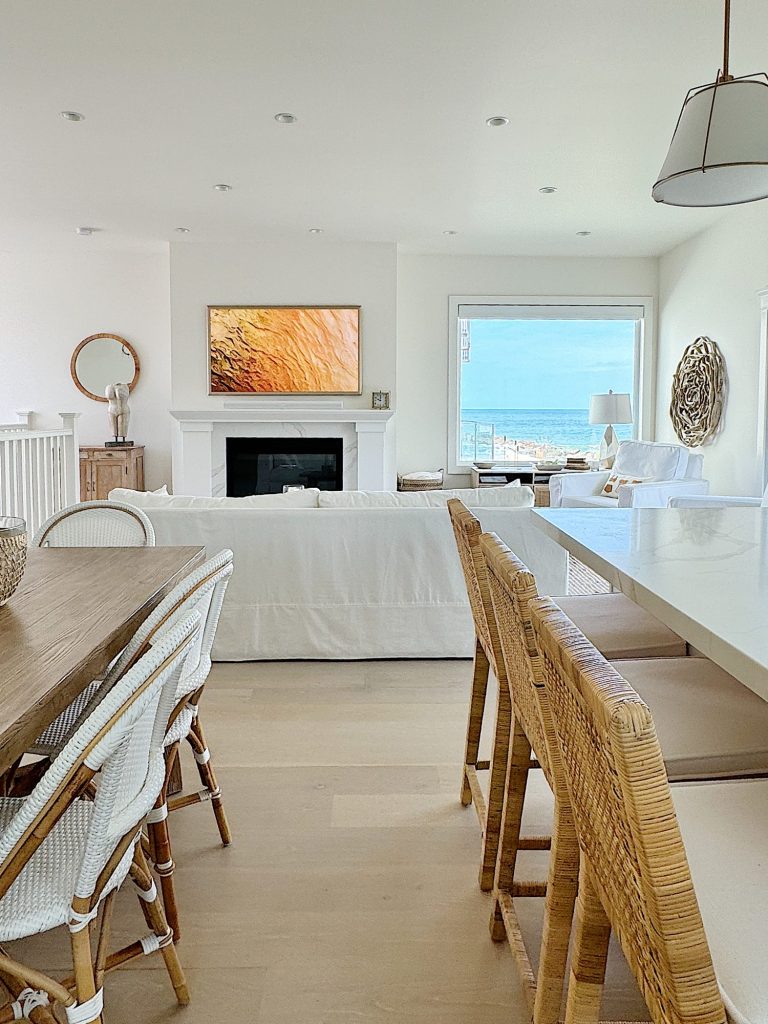 Bright, modern living room with an ocean view, featuring a wooden dining table, wicker chairs, a white sofa, and abstract art above a fireplace.