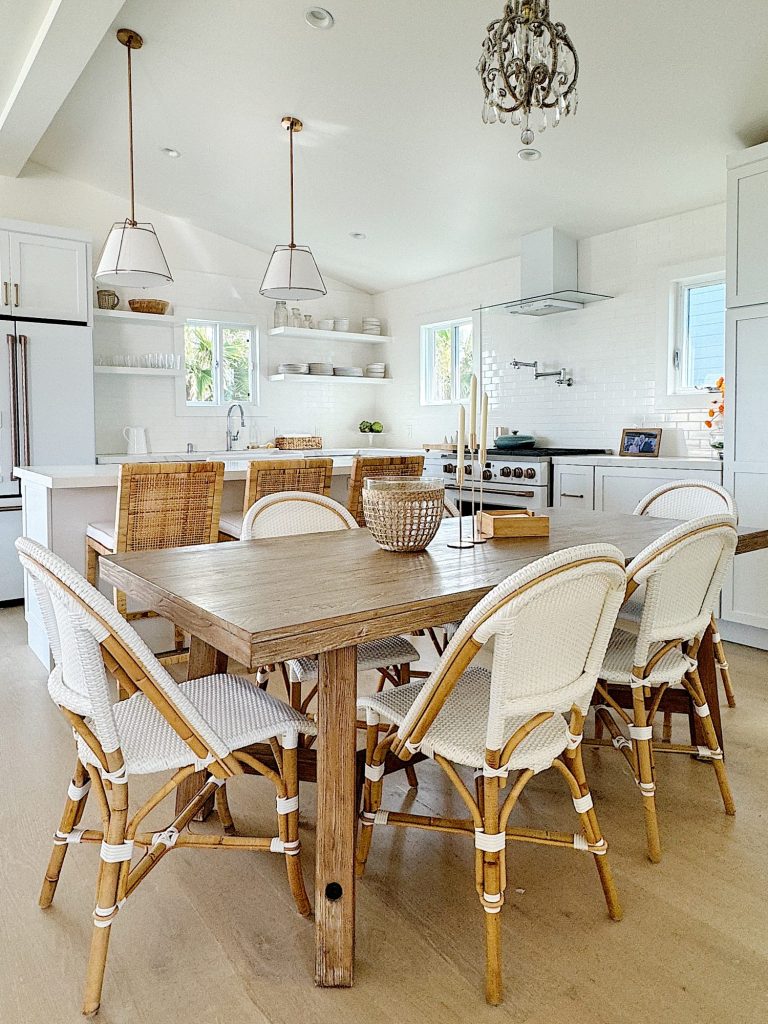 Bright kitchen with a wooden dining table, wicker chairs, white cabinetry, pendant lights, and a chandelier, reflecting a modern farmhouse style.