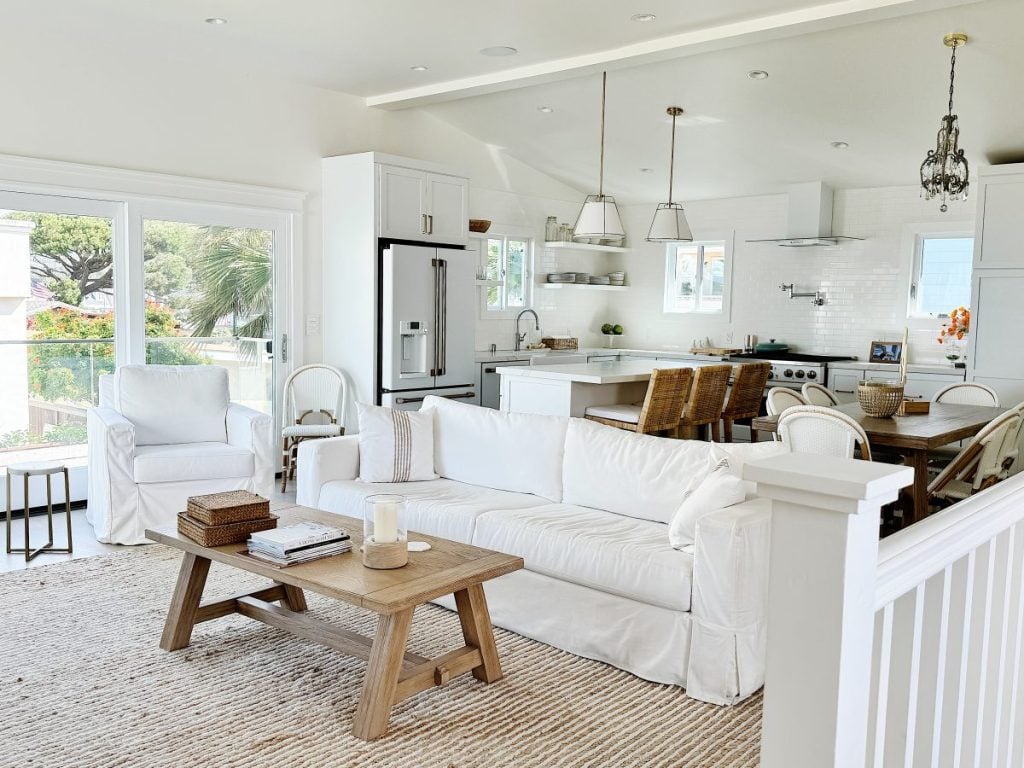 Bright, open-plan living room with white sofas, a wooden coffee table, and an adjacent kitchen with modern appliances. large windows offer a view of greenery outside.