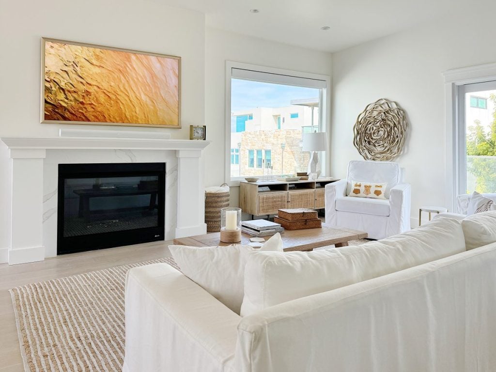 Modern living room with a white sofa, fireplace, and a large abstract painting over the mantel, with a view of another room and large windows.