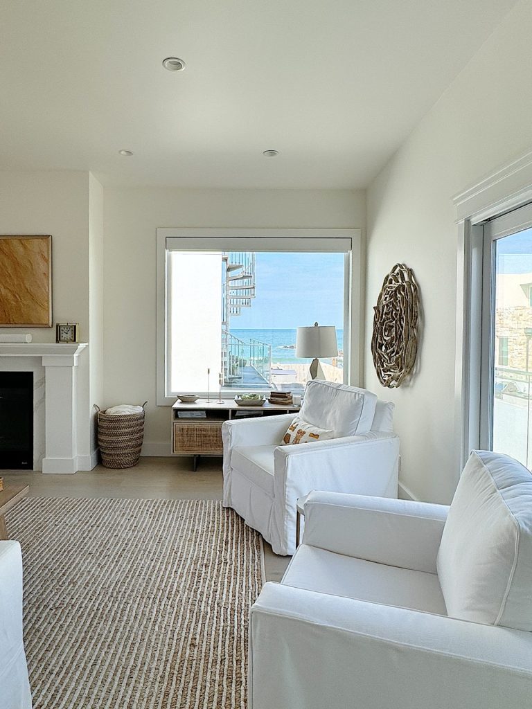 A bright living room with white sofas, a fireplace, and a large window overlooking the ocean. there is a beige rug and a seashell decor on the wall.