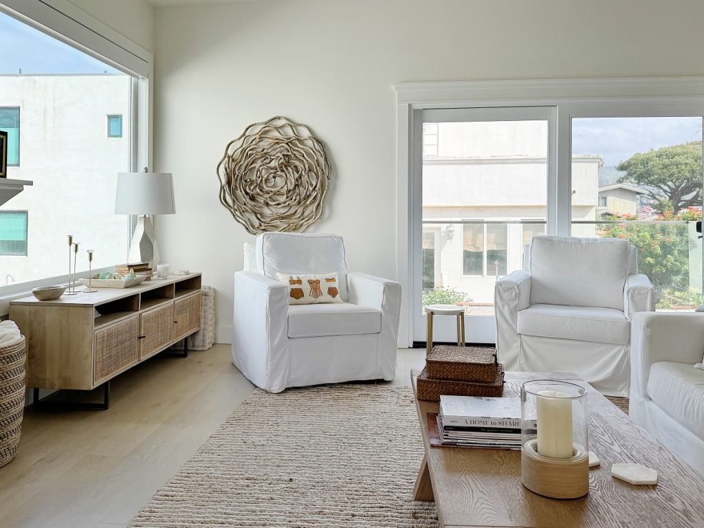 A modern, bright living room with white chairs, a decorative wall art piece, and large windows overlooking greenery.