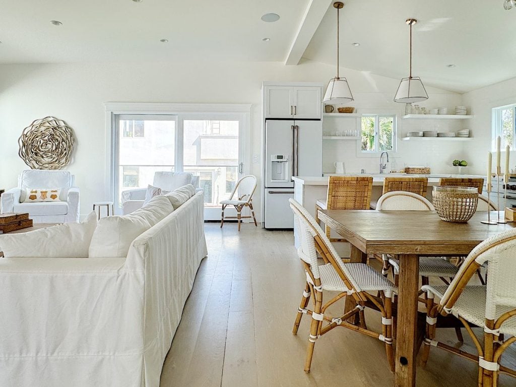 Bright, modern kitchen and living space with white furnishings, wooden table, wicker chairs, and pendant lights.