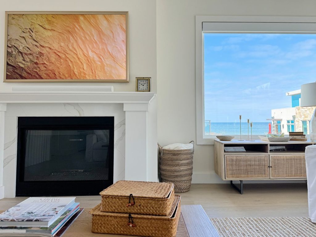 Interior of a coastal living room with a fireplace, abstract art above, wicker baskets, and a view of the ocean through a large window.