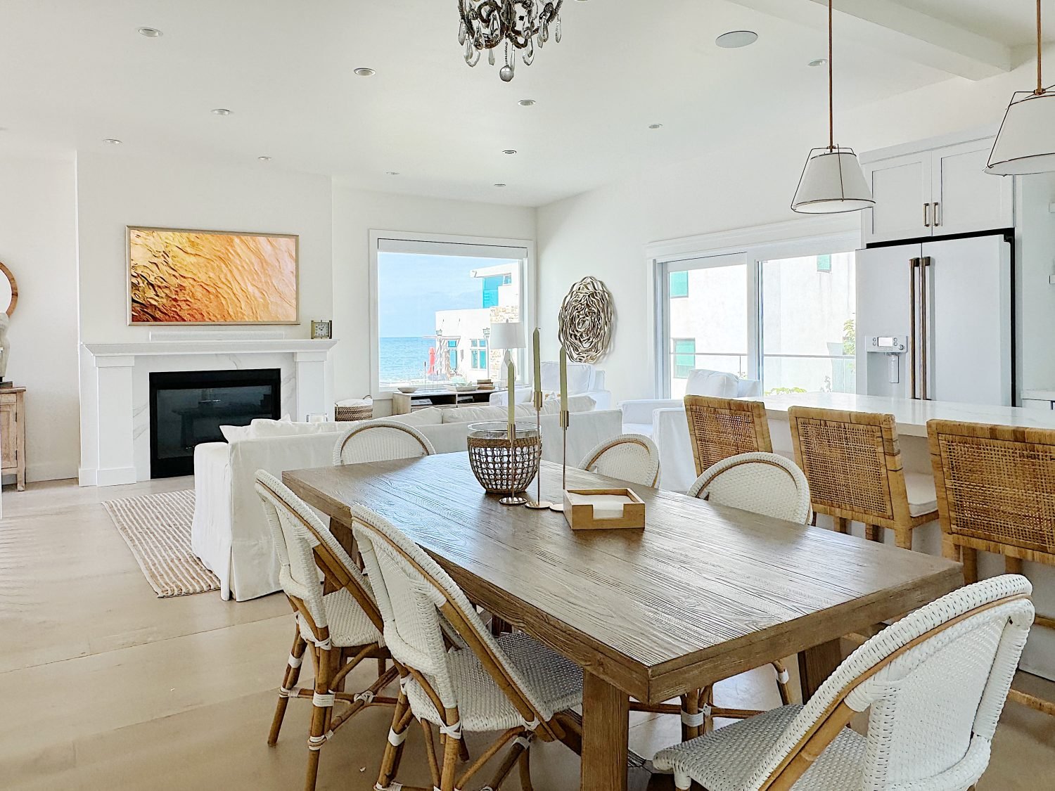 Bright, modern living room with a dining area, featuring a wooden table, white chairs, a fireplace, and a large screen displaying art.