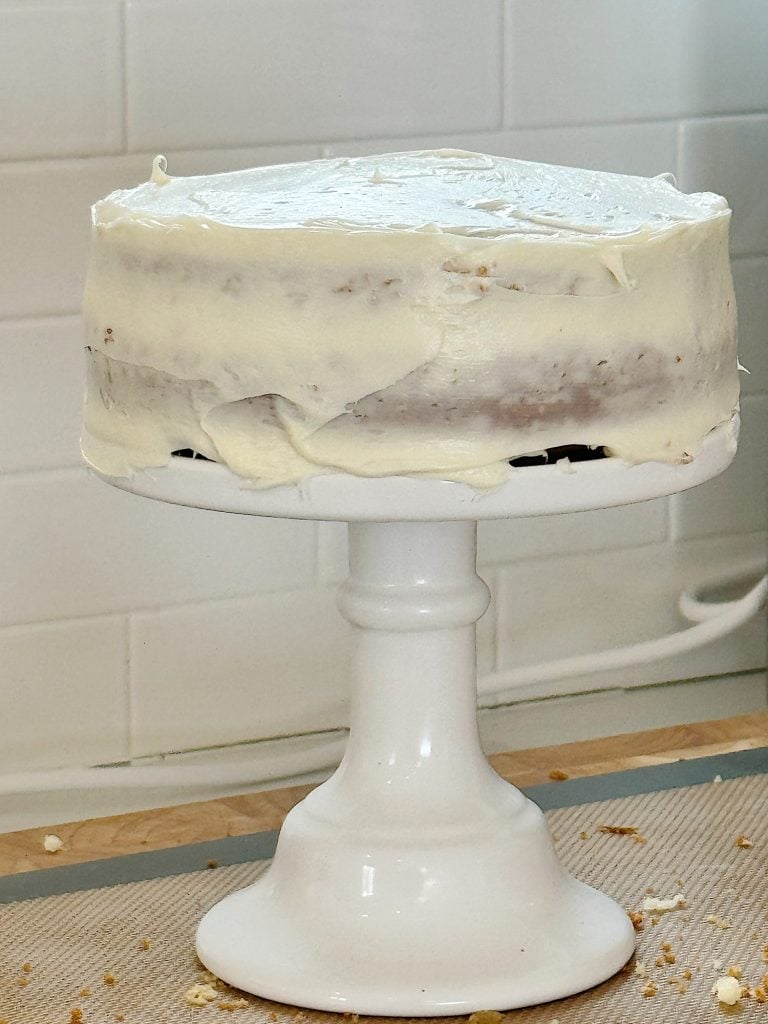 A frosted cake with white icing sits on a white pedestal stand, with some crumbs on the countertop below.