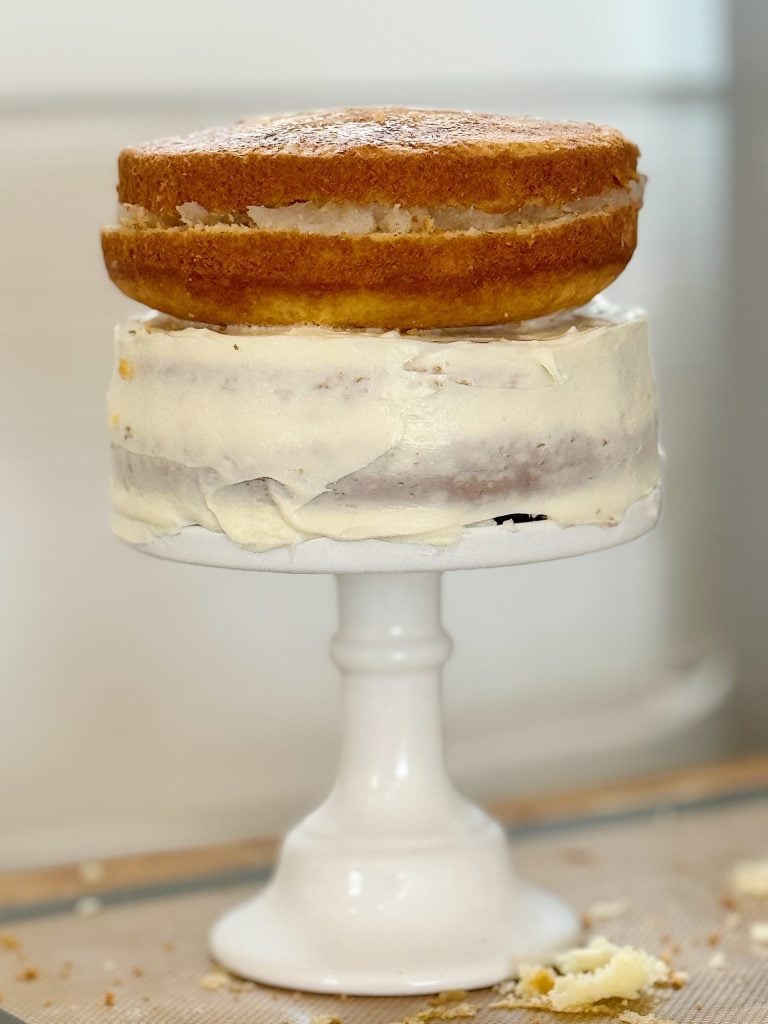 A partially frosted, two-tiered cake on a white cake stand. The top tier is unfrosted with a single layer, while the bottom tier has a thin, uneven layer of white frosting.