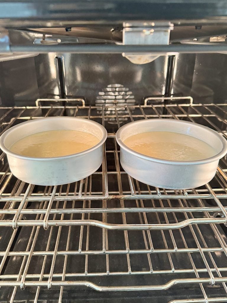 Two round cake pans filled with batter sit side by side on an oven rack inside a preheated oven.