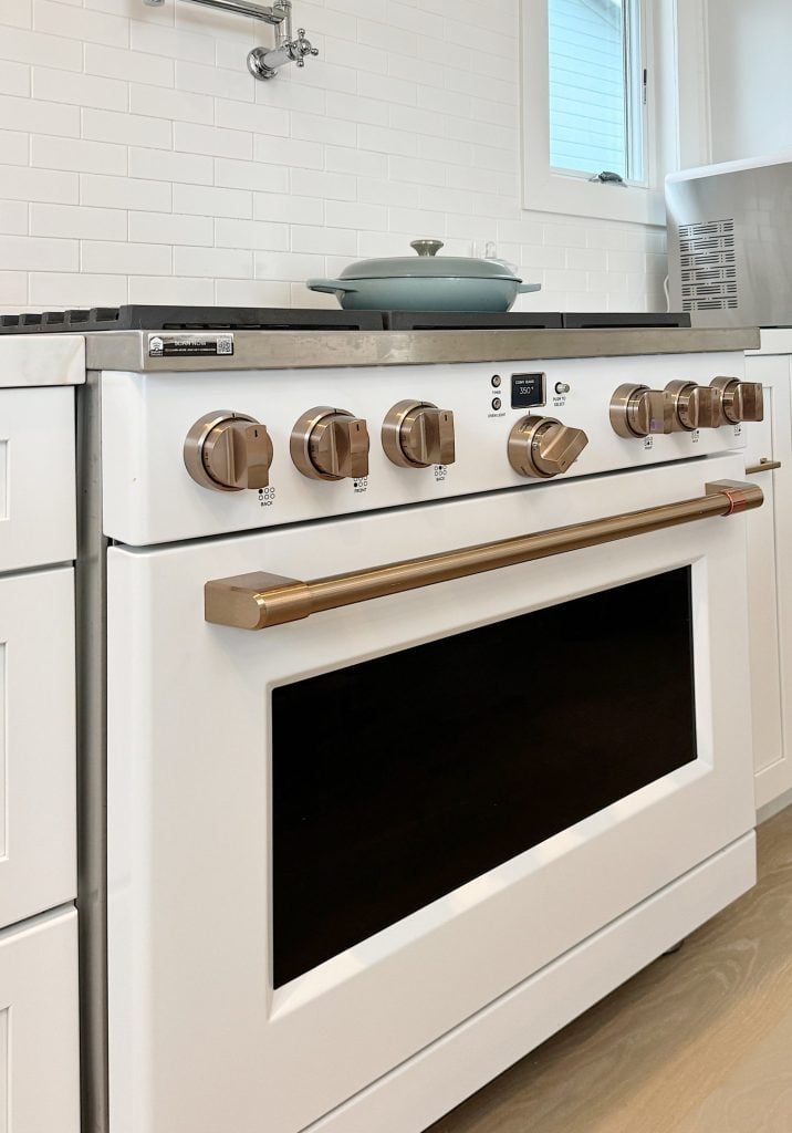Close-up of a white kitchen stove with six golden knobs and a green pot on the cooktop. The backsplash features white subway tiles and there is a small window in the background.
