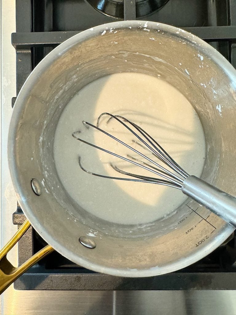 A metal whisk rests in a saucepan containing a white liquid, positioned on a stovetop burner.