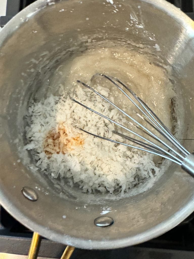 A metal pot on a stove contains a whisk and a mixture of grated coconut, flour, and a brown liquid.